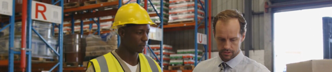 A man wearing a safety vest and hard hat takes notes as another man dressed in a shirt and tie operates a laptop.