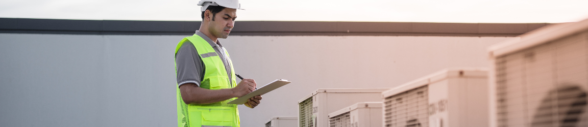 A professional in safety gear, including a helmet and reflective vest, is inspecting or recording details on a clipboard amidst a row of outdoor HVAC units during sunset. The sky is painted with the soft hues of sunset, adding a serene backdrop to the diligent work being performed.
