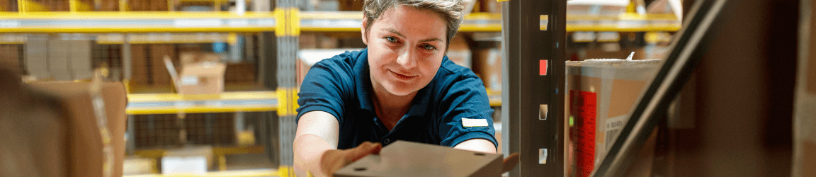 In a warehouse, a woman facing the camera wears a navy shirt and places a box on a nearly full metal shelf.