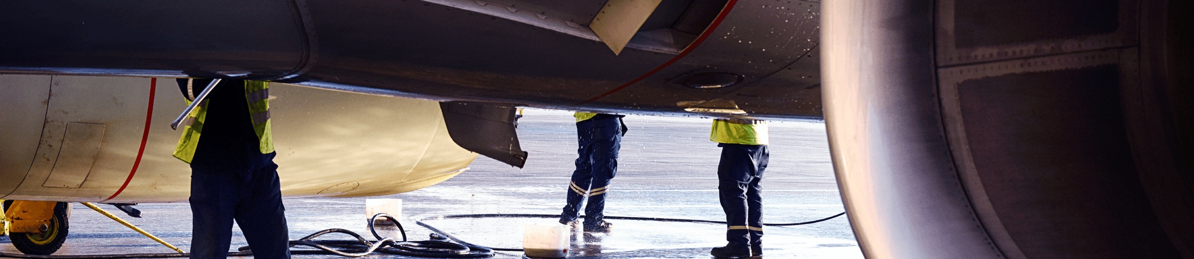 Two workers in high visibility vests are conducting maintenance on the underside of a large airplane, focusing on its engines and landing gear. Maintenance equipment is visible on the wet ground, indicating active work. The scene is dimly lit with some natural light.