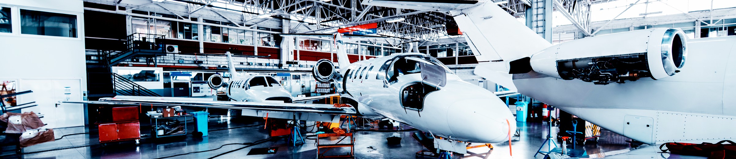 The brightly lit aircraft maintenance hangar features two white jets. In the foreground, a jet with an open nose cone reveals internal components. Behind it, another intact jet awaits service. Tools and equipment are scattered on the floor, and a staircase leads to an upper level.
