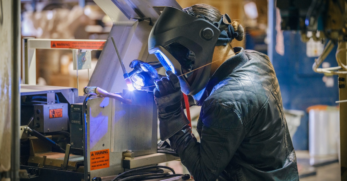 In an industrial environment a welder wearing a welding masking and heavy protective garments welds while producing a bright blue flame. 