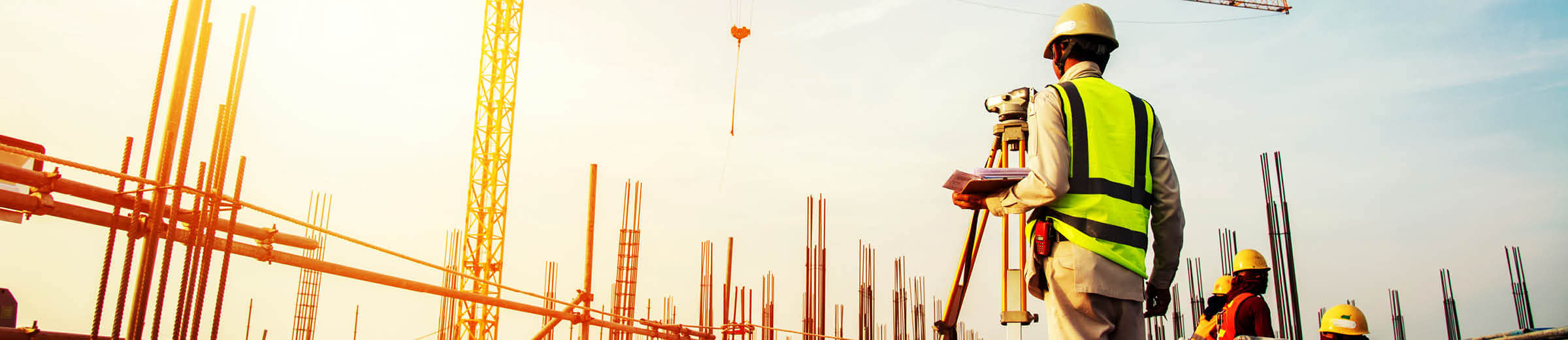 A worker in a yellow vest and hard hat looks up, beyond a surveying tool, at a worksite