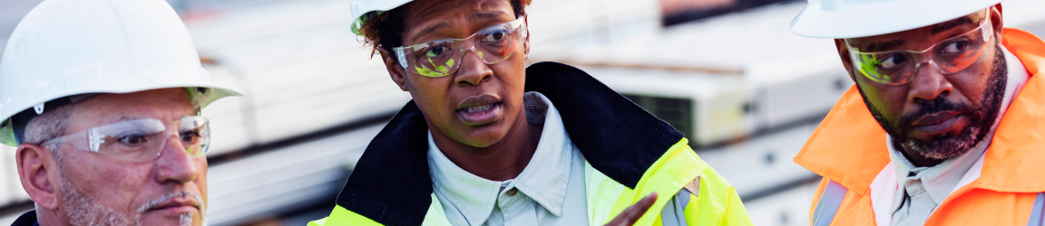 A confident construction foreman stands at a construction site wearing a white hard hat, safety goggles and a reflective vest. They hold a digital tablet and appears to be overseeing the site, suggesting their role as a supervisor or foreman. The background includes construction equipment and materials, emphasizing an active work environment.