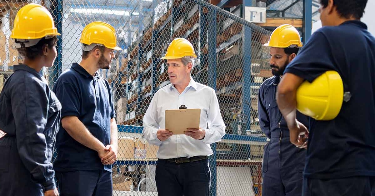 A team of preventative maintenance technicians wearing dark blue work uniforms and yellow hard hats gather to discuss the opening of a new automated food distribution center. 