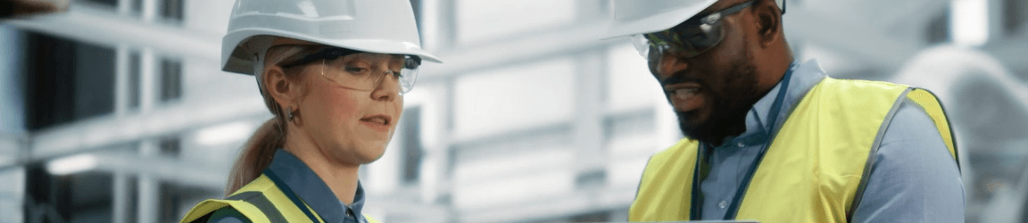 On a construction site, a woman on the left and man on the right are standing while wearing hard hats, safety glasses, yellow safety vests and grey shirts. Together they review data on a laptop computer.