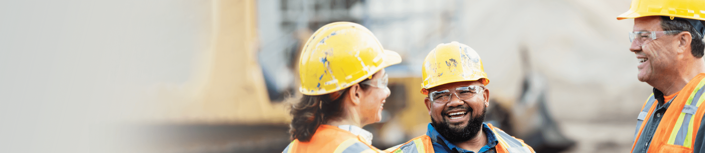 Three construction workers wearing yellow hardhats and orange vests talk to eachother on a job site.