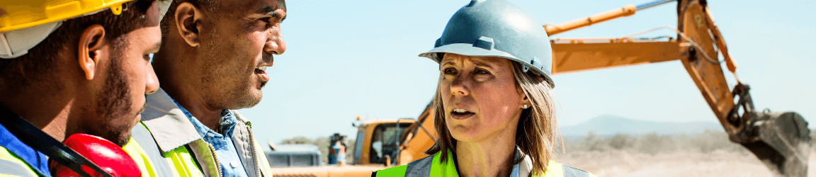 On a construction site, a woman wearing a grey safety hat and safety vests talks to two men wearing similar attire. In the background, an excavator drops dirt into a truck.
