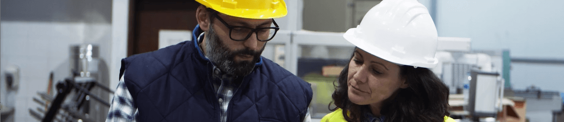 A man in glasses wearing a flannel shirt, dark vest and hard hat shows a woman in a bright yellow coat and hard hat information on a tablet.