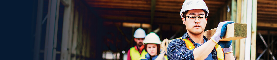 Three workers in hard hats carry a beam at a construction site