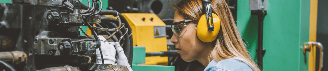 In a factory we focus on the side view portrait of young female technical repair employee wearing ear protection, safety gloves and a blue work shirt. She is operating a production line industrial CNC machine and testing, installing, analyzing and fixing bolts it with hand tools for work at a modern factory plant building.