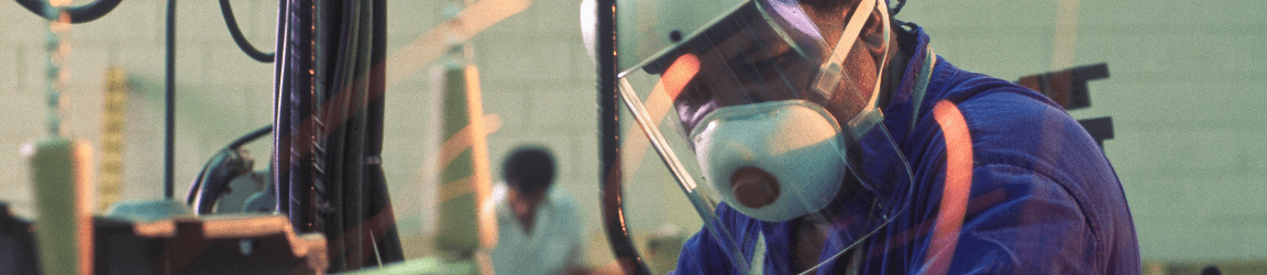 A worker wearing heavy protective clothing, face shield and respirator operates machinery in an automobile factory.