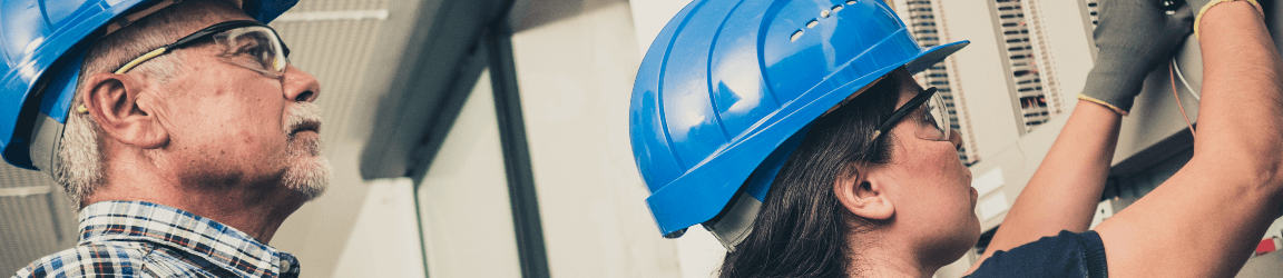 In a small room, a man and a woman wearing blue hard hats work on an electrical box on a wall.
