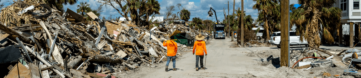 On a sunny day, two workers walk away from down a dirt covered road facing away from the camera. They are wearing orange long-sleeve shirts, blue denim pants and work boots. To their left, piles of debris line the street. To their right, several homes and cars remain intact. Aerotek can help companies overcome staffing challenges in disaster affected areas.