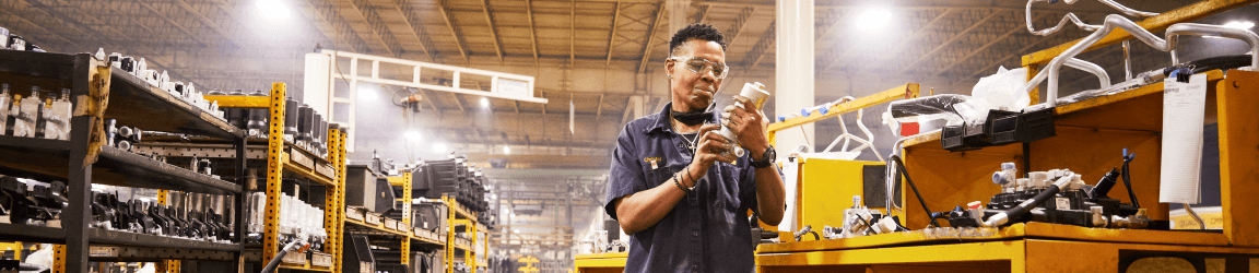 A woman wearing dark work clothes and safety glasses closely inspects a replacement component. 