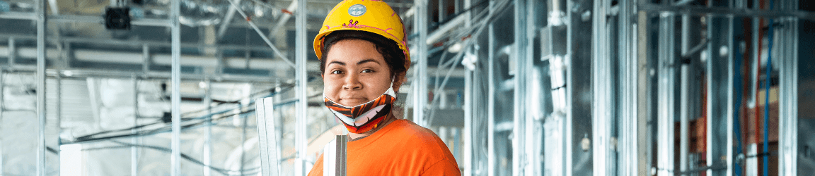 A woman constructing the interior of a building wears an orange shirt, hardhat and tool bag while holding a metal beam and smiling at the camera.