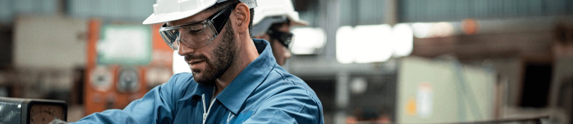 A man in a hard hat and safety glasses looks down at what he is working on in a warehouse environment.