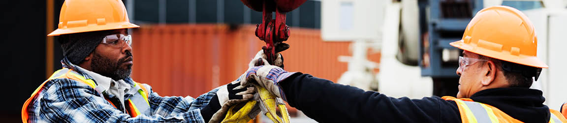 Two construction workers in orange hard hats reach for the same hook in the middle of the frame.