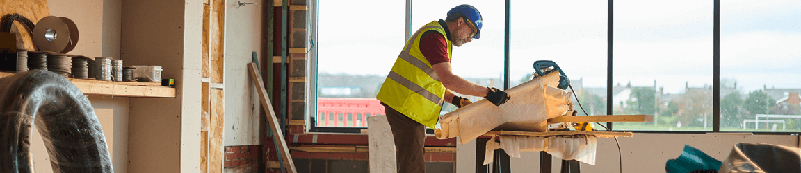 A worker wearing a reflective vest and hardhat uses a saw to cut wood in a building.