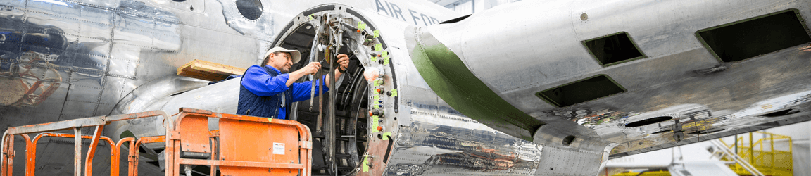 A worker in a blue vest and hat examines a part of a plane engine on a silver plane inside a warehouse.