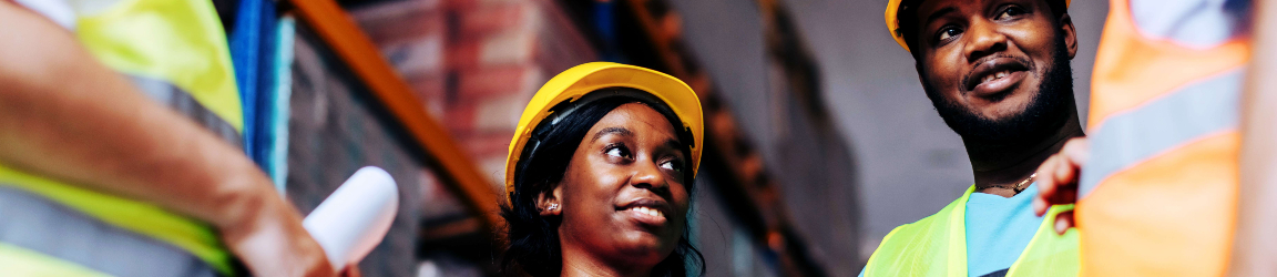 A group of warehouse workers wearing safety vests and hard hats stand together in an industrial distribution setting, reviewing documents and a tablet during a worksite discussion.