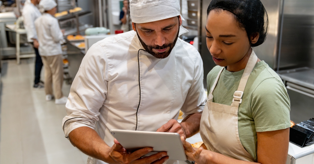 Two food service employees, wearing aprons and standing behind a counter, look at a digital tablet together. The counter is filled with baking ingredients and utensils, suggesting they are following a recipe. The setting is a bright, modern industrial kitchen with shelves and baking tools in the background.