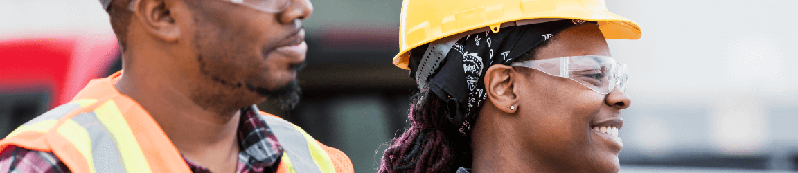 On a construction site, a woman smiles while wearing an orange safety hat, safety glasses and a high visibility vest over a flannel shirt. She is walking next to a man wearing a white hard hat, safety glasses and a high visibility vest over a flannel shirt.