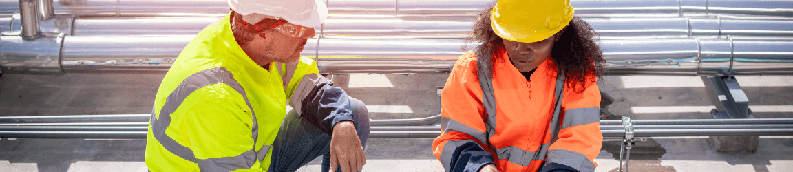 On a roof, a man and woman wear yellow safety jackets while kneeling over several metallic pipes. On the left, the man wears a white hardhat and faces the kneeling woman wearing an orange hardhat on the right. Aerotek can help you find the right HVAC technician job.