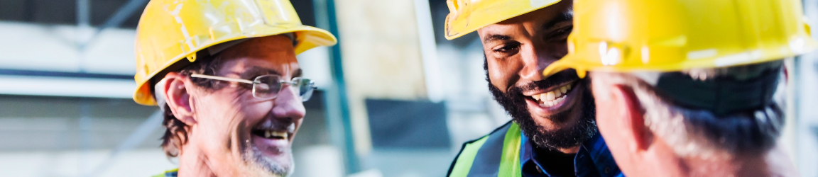 Three construction workers wearing safety vests and hard hats, standing together and smiling at a worksite.