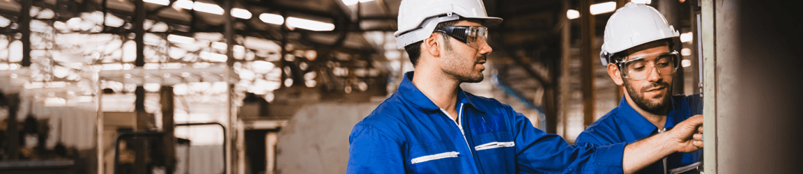Two workers wearing blue shirts, hard hats and protective glasses examine a wall.