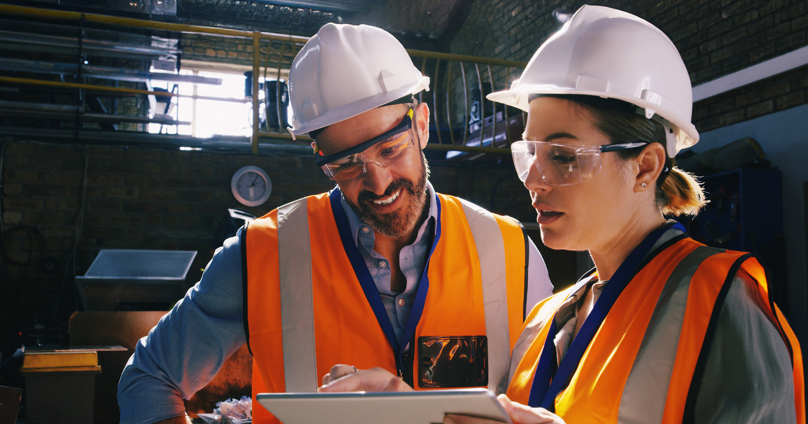 Manufacturing supervisors wearing hard hats and high-visibility vests collaborate using a tablet on an industrial worksite.