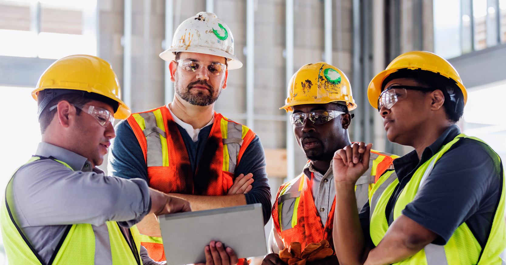 Construction professionals wearing hard hats and safety vests examine information on a tablet during an onsite project discussion inside a commercial building.
