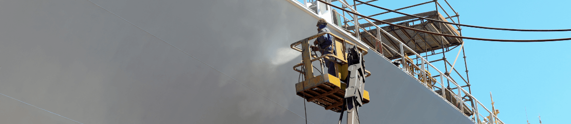 A worker paints the side of a large military style ship safely from a raised platform.