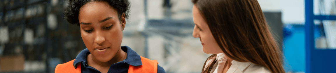 A female worker in an orange vest talks with another female in a white collared shirt.