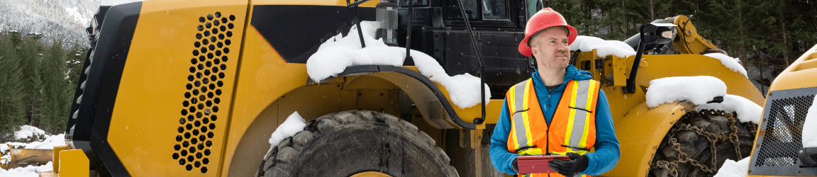 In a snowy remote location, a man stands in front of a bulldozer while wearing a safety hat, vest, blue hooded sweatshirt and blue jeans. There are mountains and pine trees in the background.