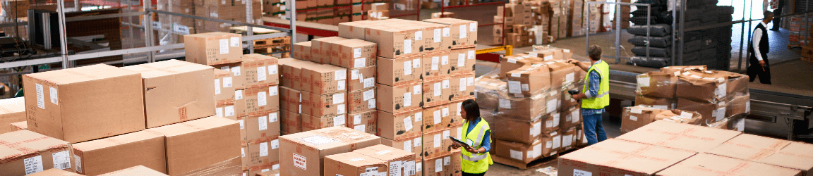 Three workers wearing safety vests organize dozens of boxes for shipment inside of a warehouse. 
