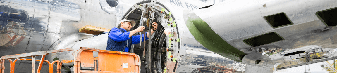 A Caucasian male aviation mechanic on a mechanical lift, works on an airplane engine in an airplane hangar. He wears a blue shirt, dark vest and white baseball cap.