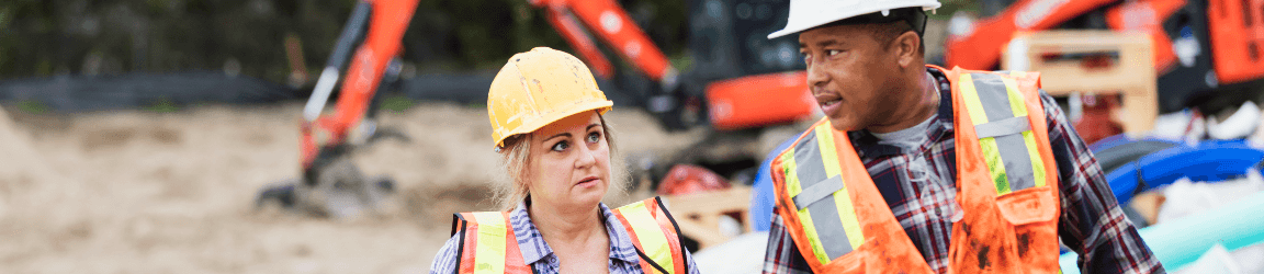 Un homme et une femme portant des casques et des gilets protectifs marchent à travers un chantier. 