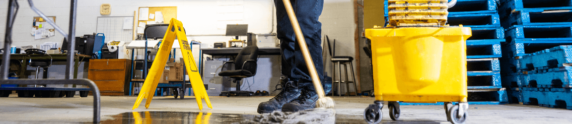 Un homme portant des lunettes de sécurité, des gants de protection, une chemise grise et un pantalon foncé utilise une vadrouille et un seau pour nettoyer un dégât liquide dans une installation de fabrication. Il a posé un panneau d’avertissement à proximité du dégât.
