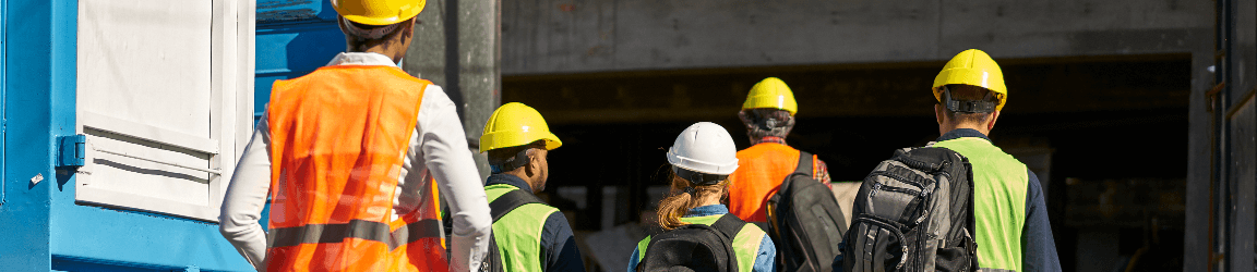 A rear shot of a construction team walking into a building. Four workers were yellow hard hats, and one worker wears a white hard hat. All are wearing safety vests and three workers are wearing backpacks.  