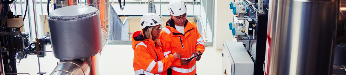  Two maintenance technicians wearing orange high-visibility jackets and white safety helmets examine a tablet together inside an industrial facility. One technician is training or upskilling the other. They stand near large stainless steel pipes and control panels, suggesting a power station or mechanical plant setting.