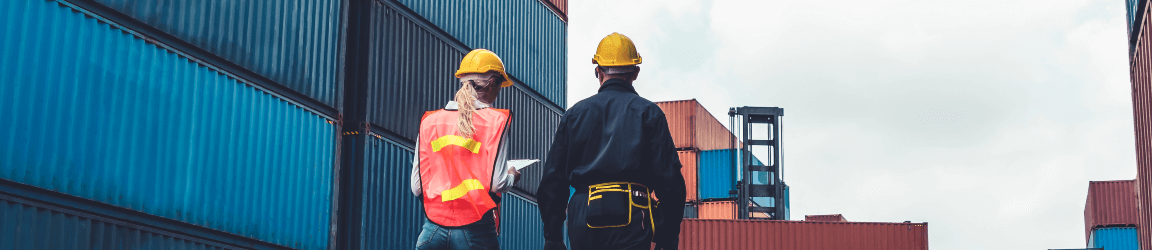 With their backs facing the camera, a man and woman wearing hard hats walk through a shipping container yard. 