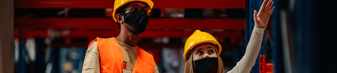 A man and woman wearing face masks, safety vests and hard hats look over the interior of a warehouse.