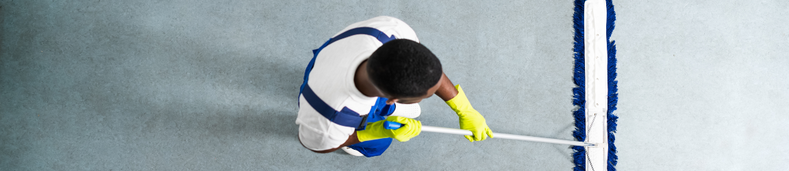 A top-down view shows a man wearing a white shirt, yellow gloves and blue overalls as he uses a large dust broom on a grey floor. The broom leaves a path of clean floor as he pulls it towards himself.