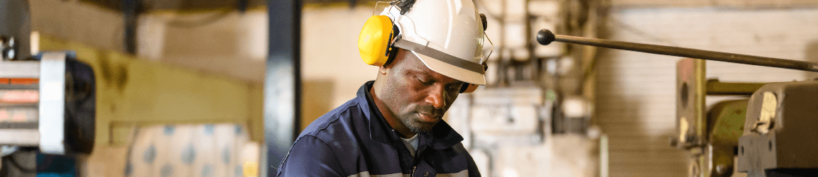A male technician in a blue shirt, wearing a hardhat and protective earmuffs, works in a shop