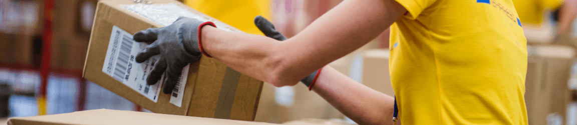 In a warehouse two men wearing blue shirts and orange safety vests use RF scanners to manage a shipment of boxes.