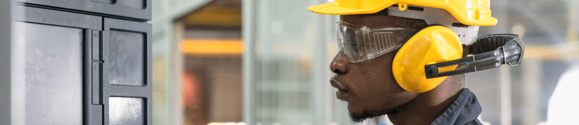 In a well-lit factory, a male control systems worker wearing dark coveralls, a bright yellow hard hat, safety glasses and bright yellow ear protection around his neck, operates industrial machinery.