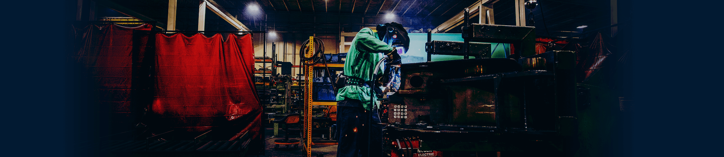 A worker in a green shirt and a protective mask welds in a manufacturing facility